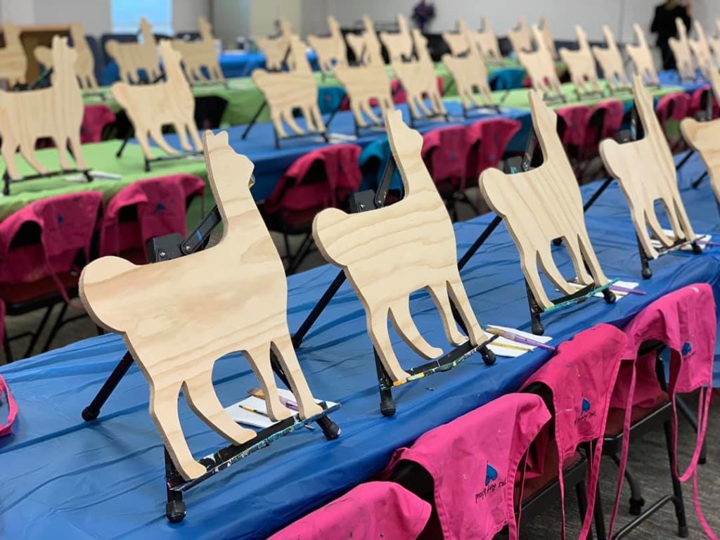 Paint party supplies - plastic table cloth, pink aprons, and rows of wooden llamas ready to be painted